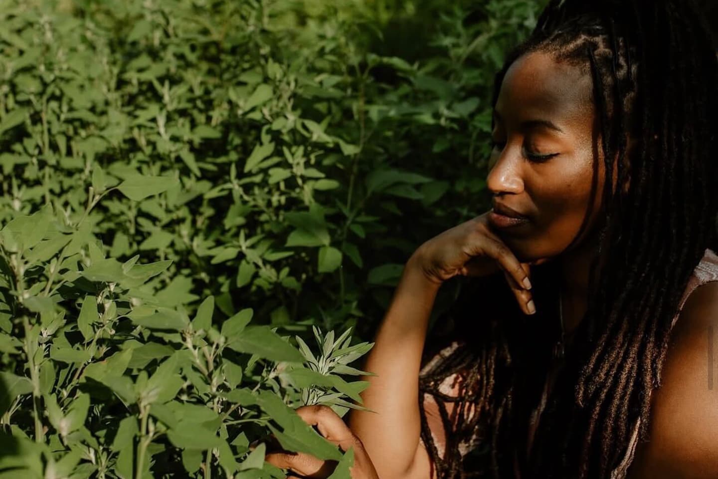 Woman with long dreadlocks seated among green plants, resting her chin on her hand while touching a leaf