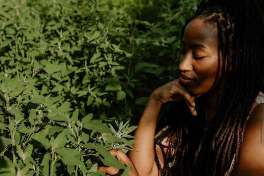 Woman with long dreadlocks seated among green plants, resting her chin on her hand while touching a leaf