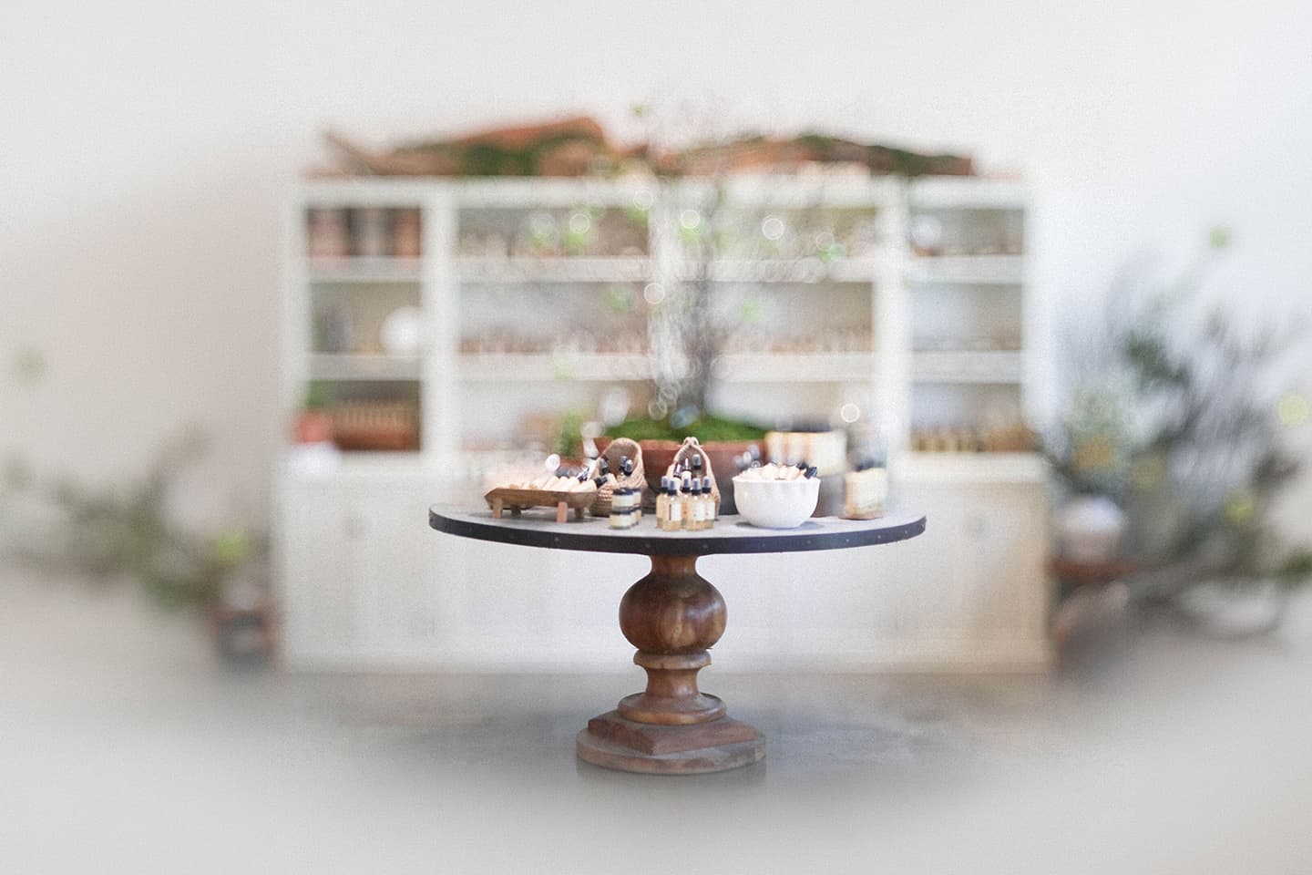 Round pedestal table displaying small glass skincare bottles, jars, and bowls in a bright boutique with blurred shelves and plants