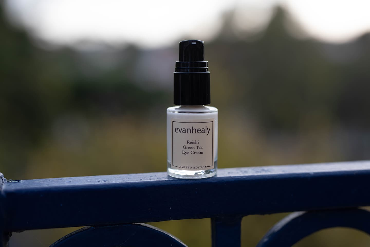 Front view of Reishi Green Tea Eye Cream bottle with black pump and evanhealy label on blue railing, blurred background