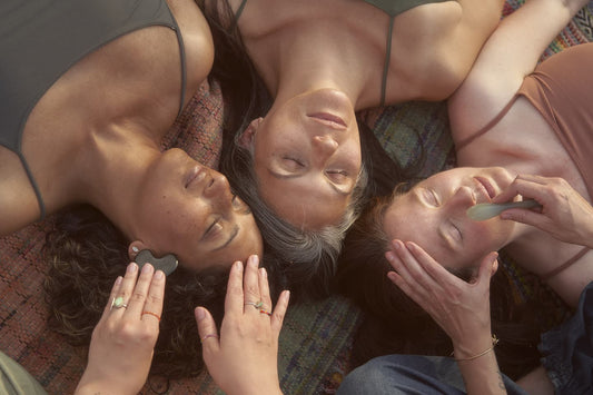 Three women lying on a patterned blanket while receiving facial massages with stone tools