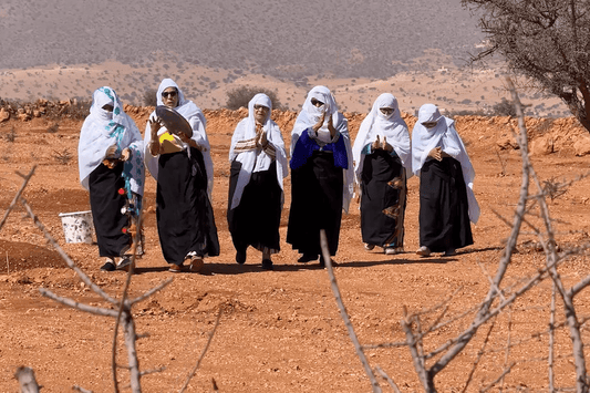 Six women in white veils and black robes stand in an arid Moroccan argan tree landscape